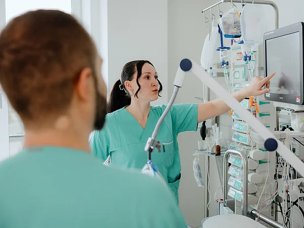 Eine Pflegekraft zeigt auf den Bildschirm eines medizinischen Geräts, während eine weitere Pflegekraft im Hintergrund zuschaut.