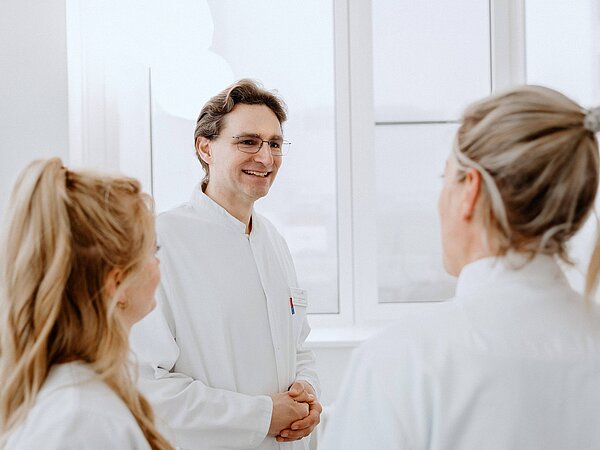 Ein Arzt steht im Gespräch mit zwei Pflegekräften, die ihm aufmerksam zuhören. Alle tragen weiße Arbeitskleidung und befinden sich in einem hellen, modernen Raum.