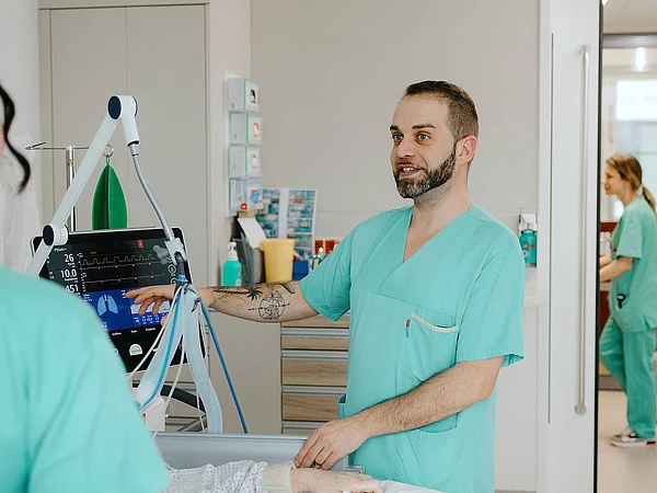 Ein Pflegekraft hält ein medizinisches Gerät und richtet es auf ein Bett. Weitere Pflegekräfte sind im Hintergrund zu sehen.“
