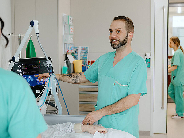 Ein Pflegekraft hält ein medizinisches Gerät und richtet es auf ein Bett. Weitere Pflegekräfte sind im Hintergrund zu sehen.“