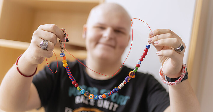 Person hält eine bunte Perlenkette, lächelt, trägt ein schwarzes T-Shirt und Ringe.