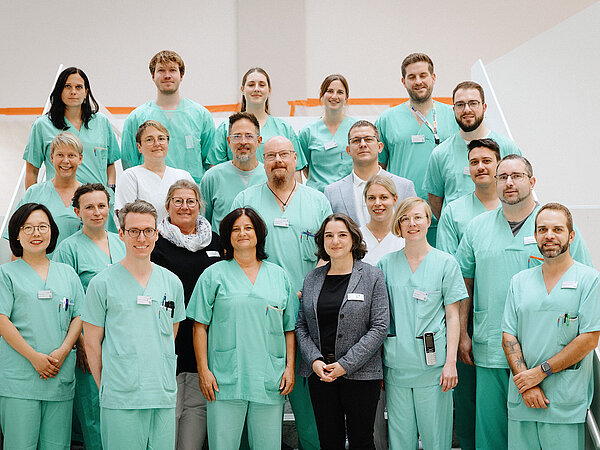 Gruppenfoto von 18 Personen in grüner Arbeitskleidung, die auf einer Treppe stehen. Die Gruppe besteht aus Ärzten und Pflegekräften, die alle freundlich in die Kamera blicken.