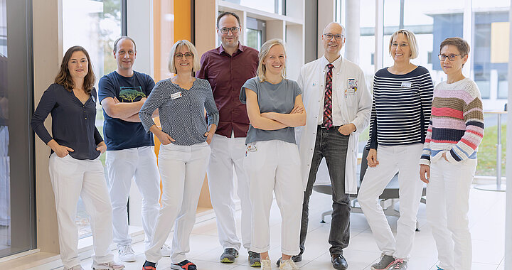 Eine Gruppe von acht Personen, darunter medizinisches Personal, steht lächelnd gemeinsam im Innenbereich an großen Fenstern.
