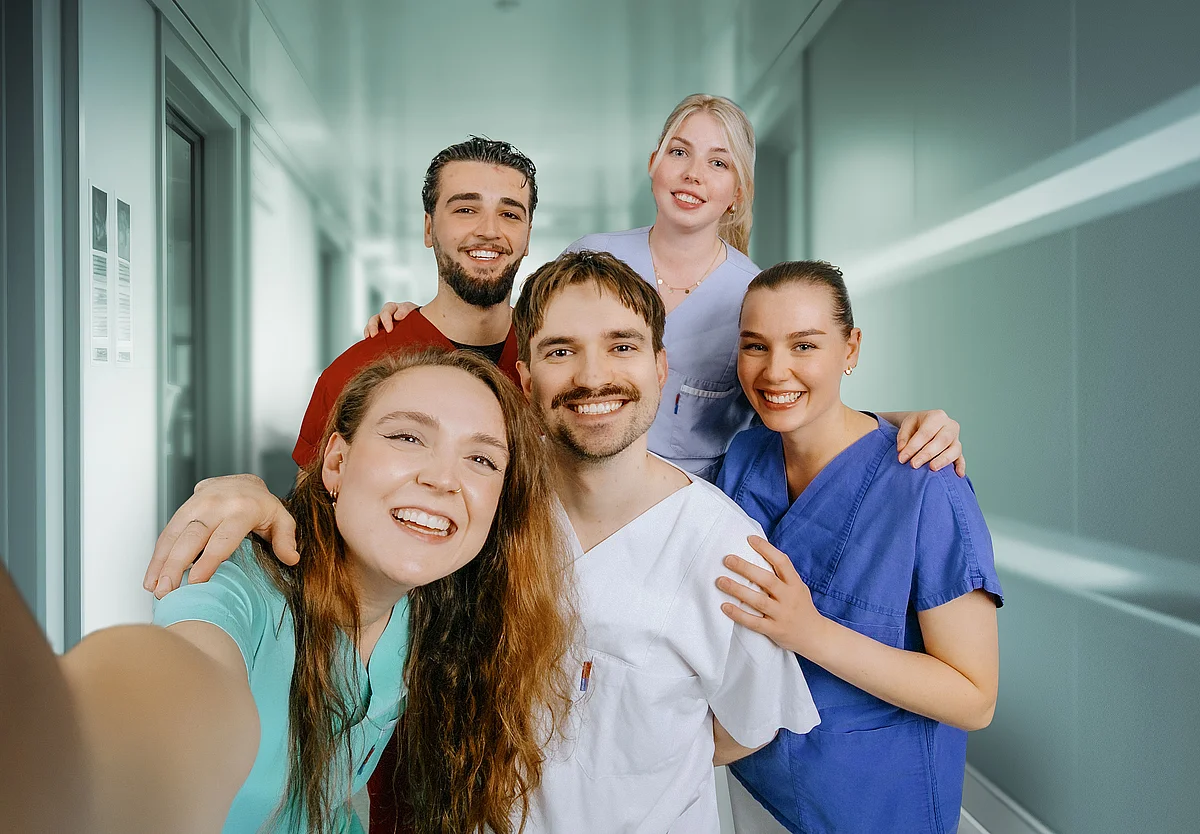 Fünf Mitarbeitende im Gesundheitswesen stehen gemeinsam auf einem Krankenhausflur und lächeln in die Kamera.