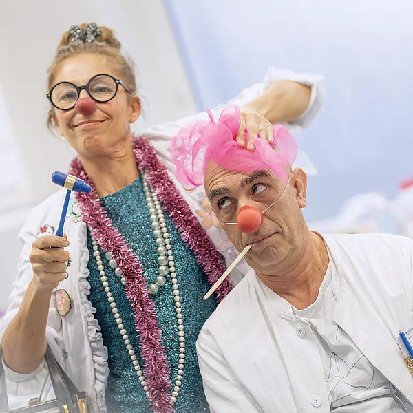 Zwei lustig als Ärzte verkleidete Personen mit Clownsnasen und albernen Accessoires posieren verspielt in einem medizinischen Umfeld.