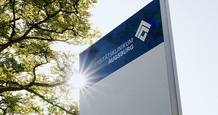 Stele des Universitätsklinikums Augsburg vor Bäumen und Sonnenlicht
