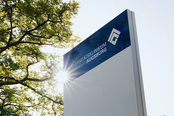 Stele des Universitätsklinikums Augsburg vor Bäumen und Sonnenlicht