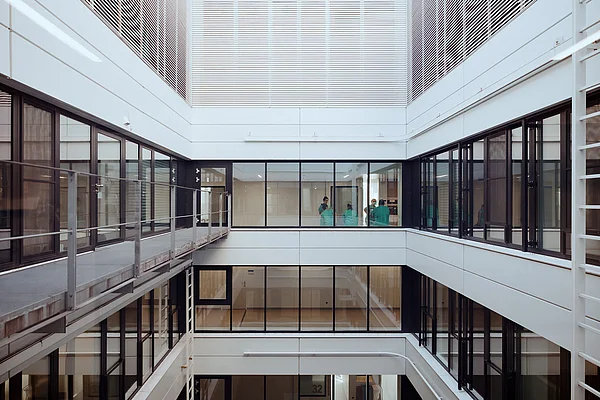 Blick auf das offene Innere eines modernen, mehrstöckigen Gebäudes mit Glaswänden und Metallrahmen. Zwei Personen in grüner Arbeitskleidung sind im Hintergrund zu sehen.