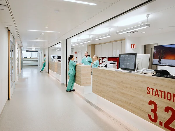 Blick auf den Empfangsbereich einer modernen Krankenhausstation mit einem Empfangstresen. Das Schild „STATION 3.2“ ist sichtbar, und Pflegekräfte arbeiten hinter dem Tresen.