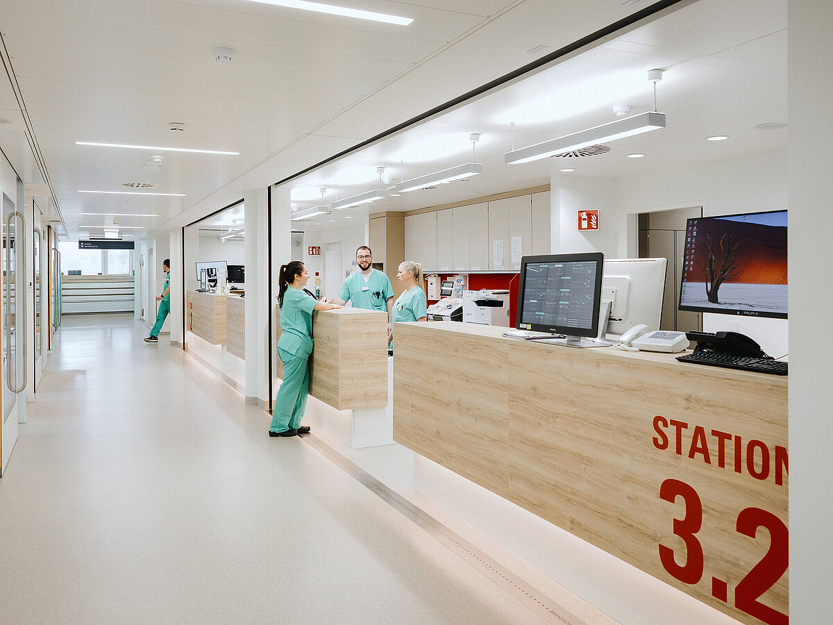 Blick auf den Empfangsbereich einer modernen Krankenhausstation mit einem Empfangstresen. Das Schild „STATION 3.2“ ist sichtbar, und Pflegekräfte arbeiten hinter dem Tresen.