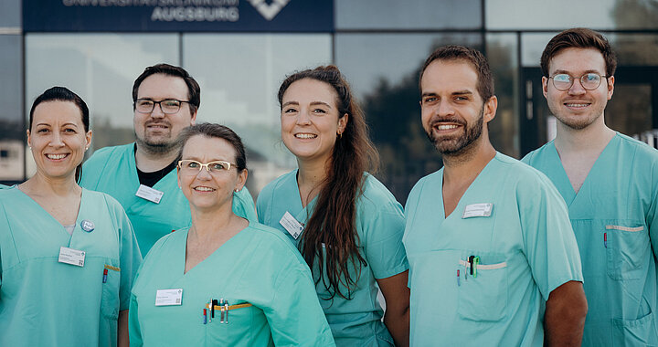 Gruppe von medizinischen Fachkräften in grünen Klinik-Kitteln vor einem Klinikgebäude.